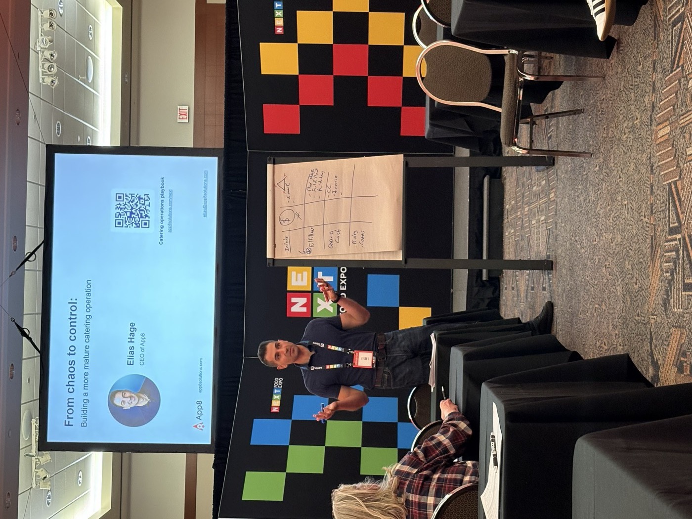 Elias Hage presenting From chaos to control at NEXT Food Expo, with the four jobs of a catering operation, intake, fulfillment, order to cash, and rules, written on a flip chart beside him.